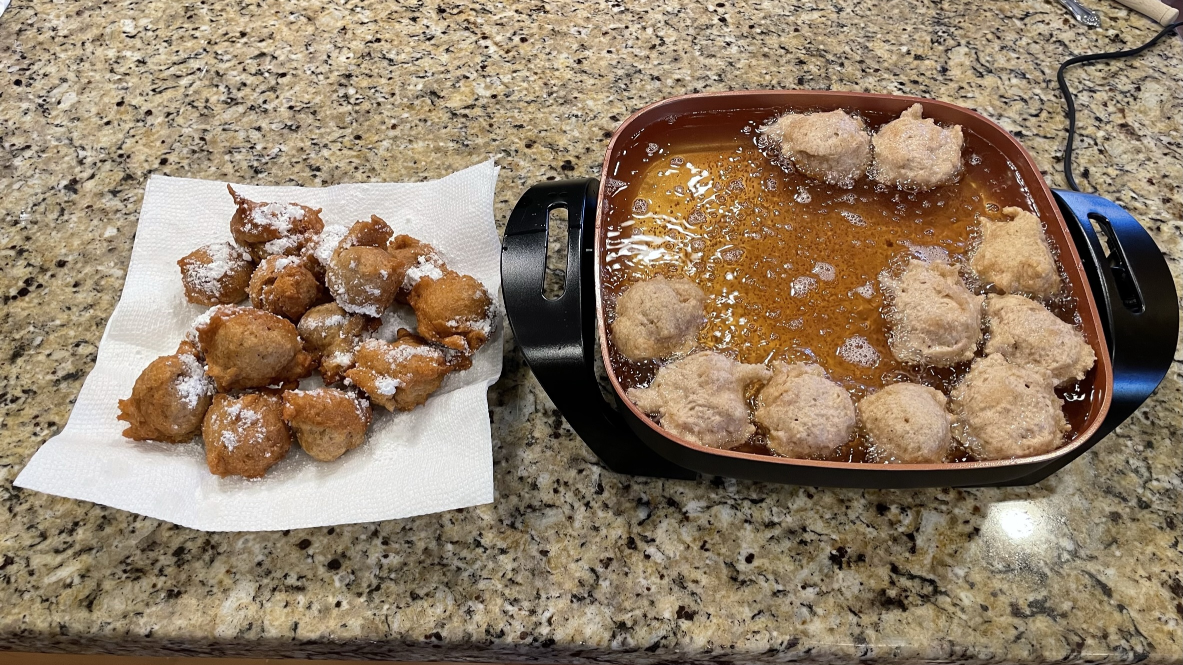 A plate of fresh Apple Cinnamon Sinkers sprinkled with powdered sugar, with more sizzling in pan of hot oil