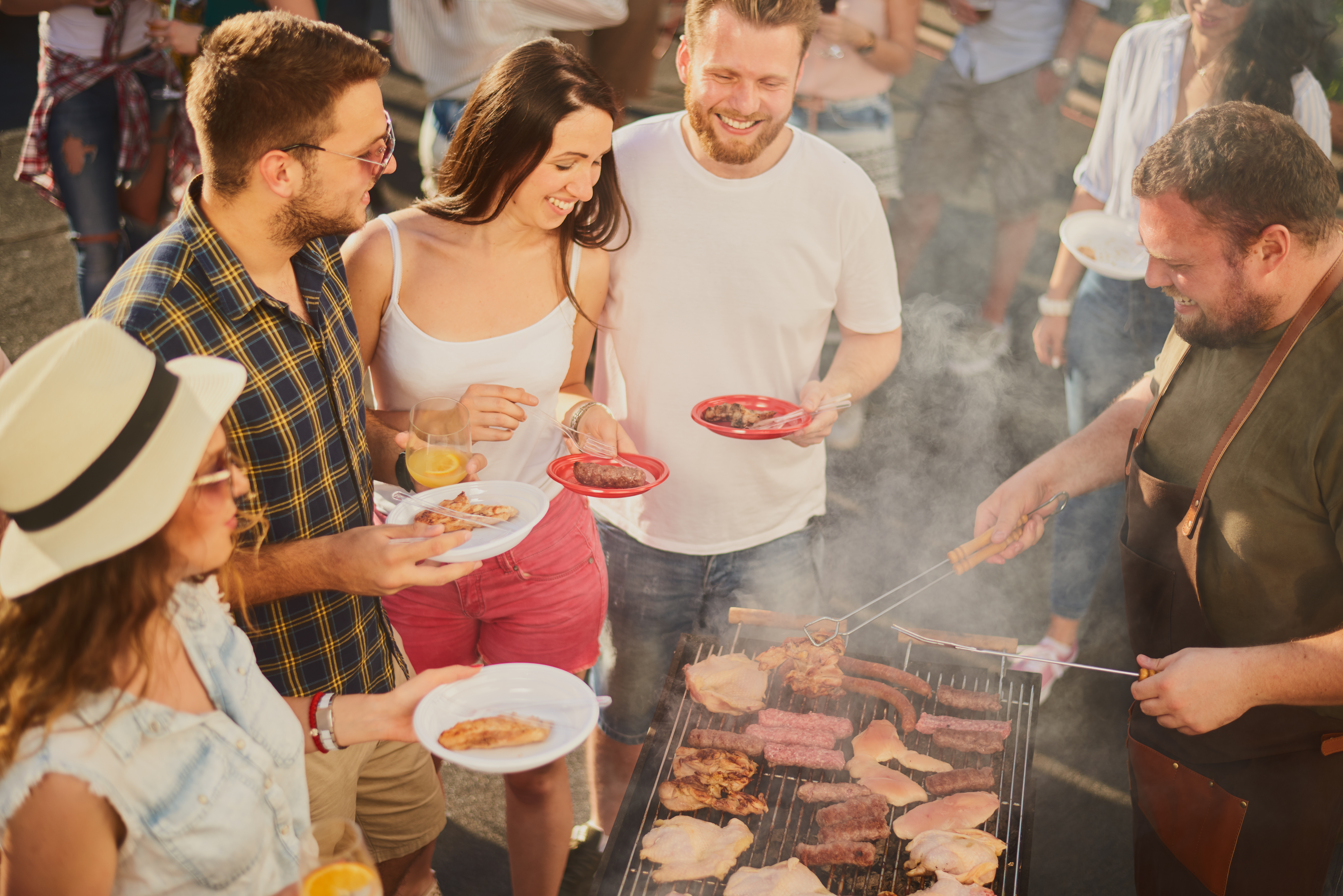 Group of people standing around grill, chatting, drinking and eating.