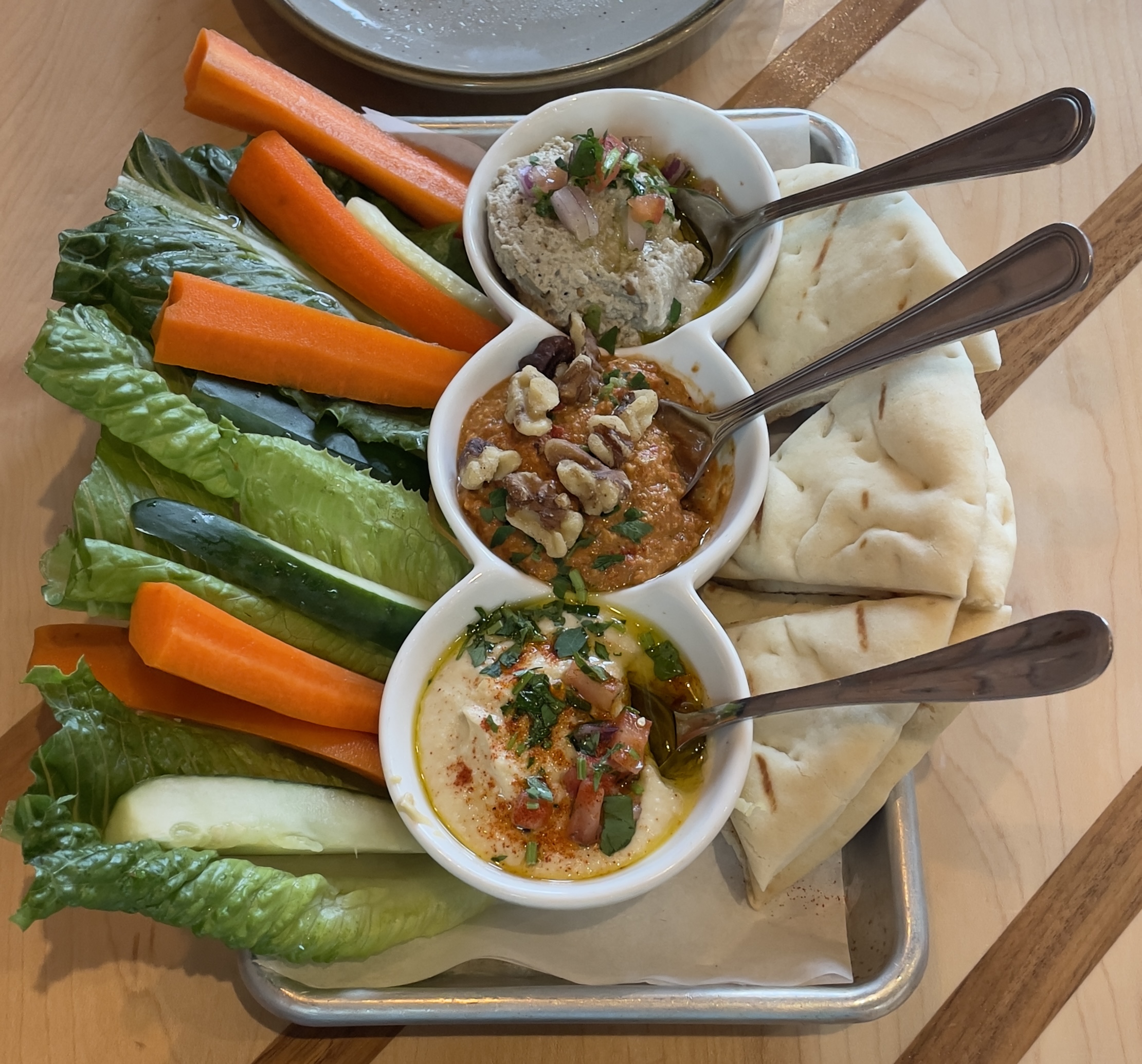 Sampler Dip Plate at Laziz Kitchen with Hummus (garbanzo beans, tahini, lemon, garlic), Muhammara (roasted red pepper locally roasted by Firebird, pomegranate molasses, onions, garlic, walnuts), and Baba Ghannouj (fire-roasted eggplant, tahini, lemon, garlic).  Served with warm pita and fresh veggies.